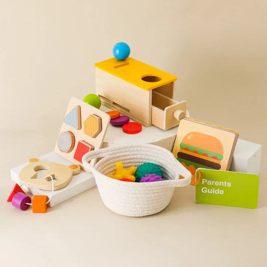 Montessori First Year Discovery Set: natural wood object permanence box with yellow top, shape and texture inset board, threading ring with chunky beads, pastel sensory balls, color discs, and woven cotton basket